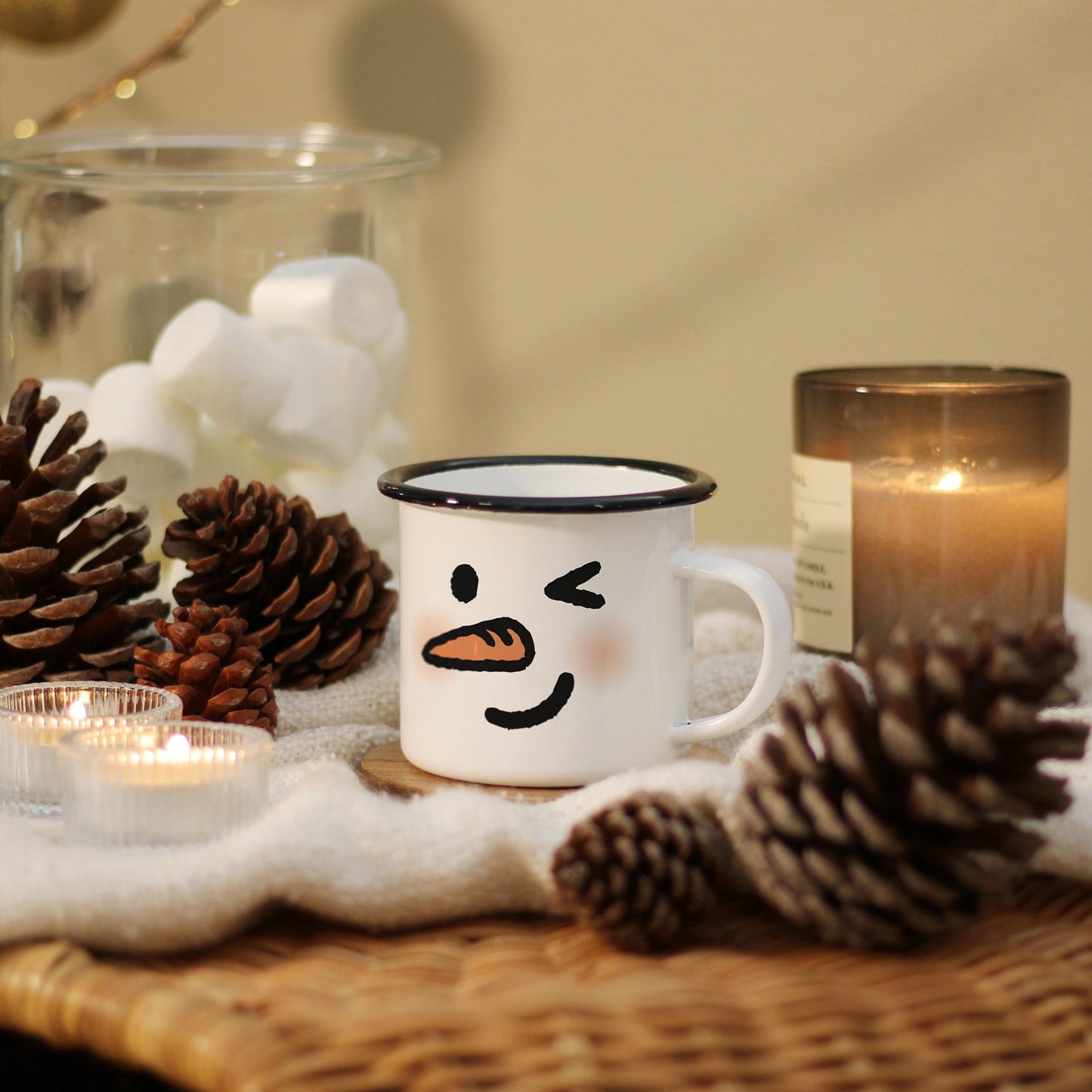 A Wink snowman mug with carrot nose and soft blushes rest on a blanket covered rattan table with pine corns and candles.