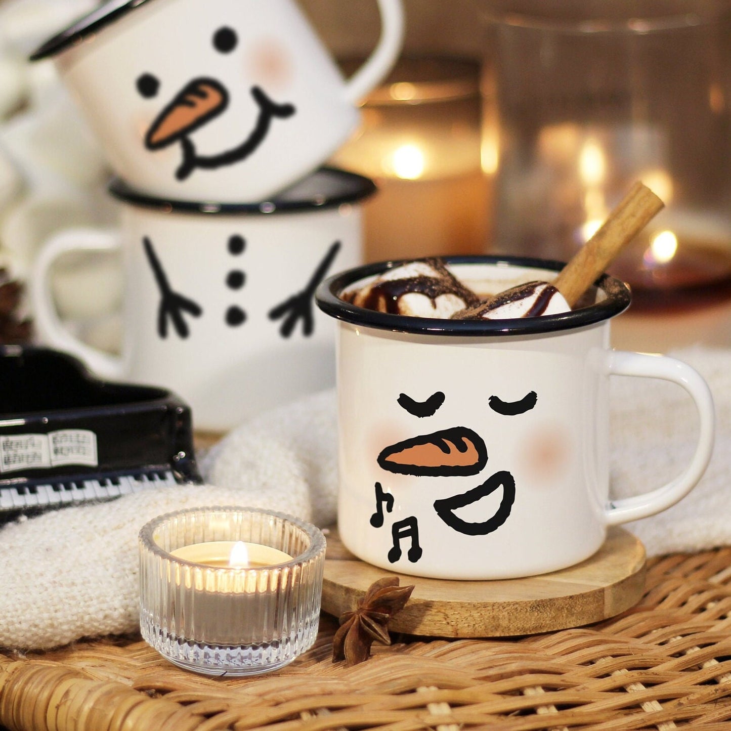 A singing snowman mug filled with hot chocolate, marshmallow, and cinnamon stick rests on a wooden coaster laid on a rattan table, surrounded by candle lights and mini piano. A mug stack of a plumped face snowman on top of a snowman body in the back.