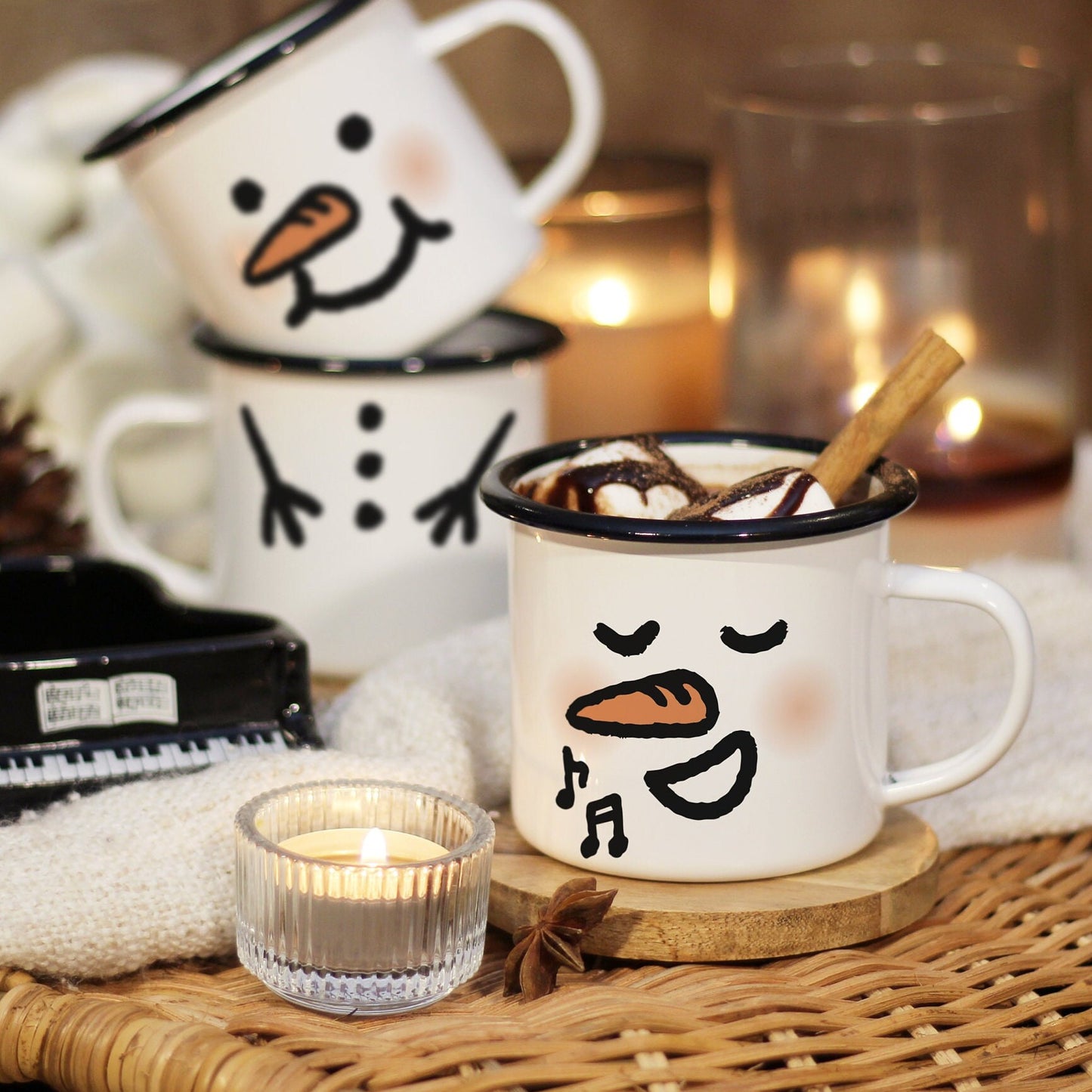 A singing snowman mug filled with hot chocolate, marshmallow, and cinnamon stick rests on a wooden coaster laid on a rattan table, surrounded by candle lights and mini piano. A mug stack of a plumped face snowman on top of a snowman body in the back.