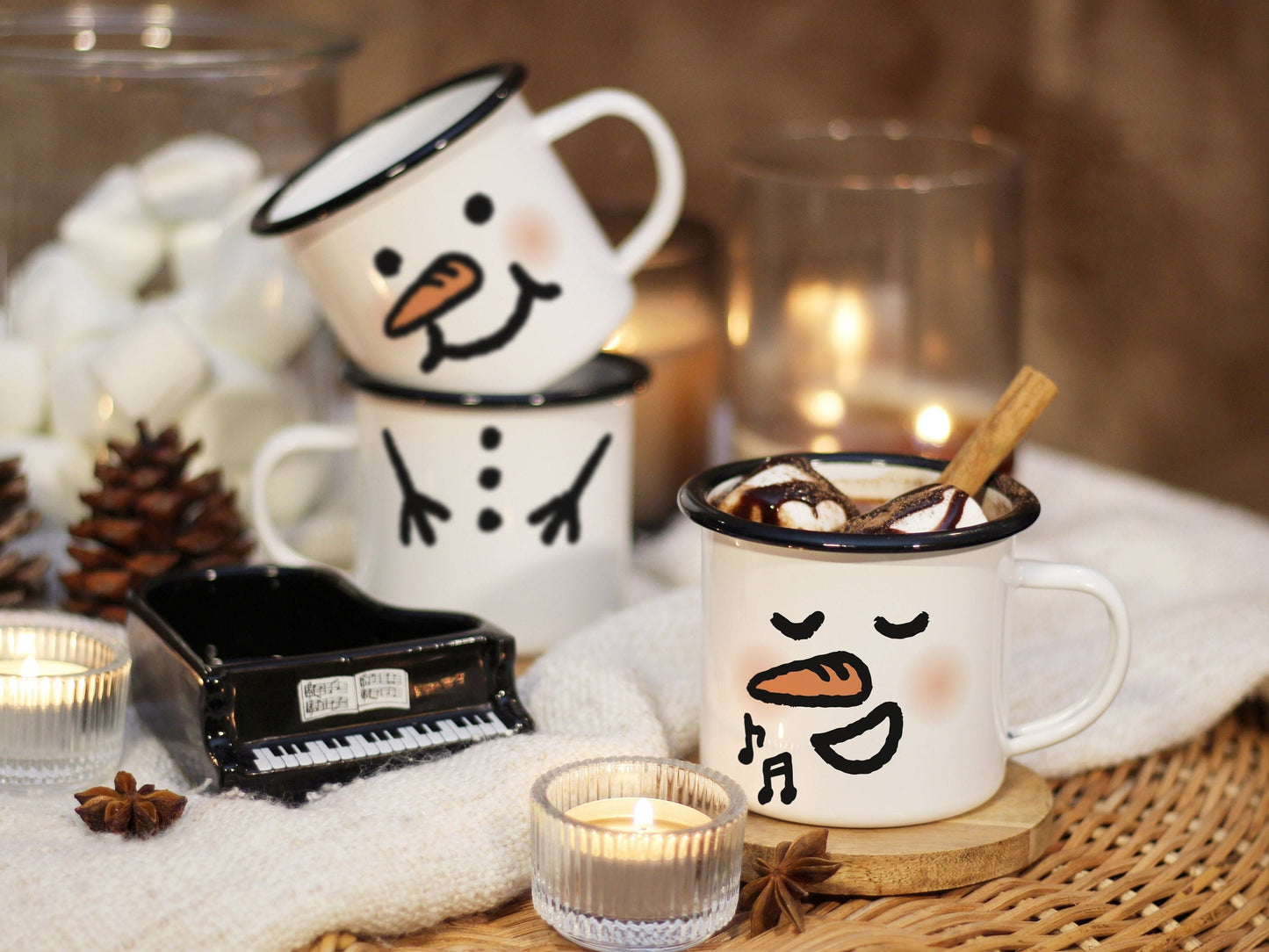 Snowman mugs on a rattan table & blanket, surrounded by candle lights, made of enamel glazed steel plate with black rim & customizable face. A singing snowman mug with hot chocolate in the front, next to a mini piano. 2 snowman mug stack in the back.