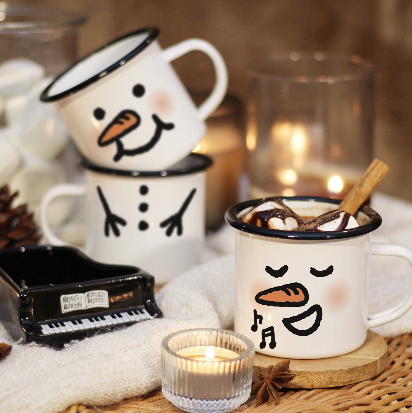 Snowman mugs on a rattan table & blanket, surrounded by candle lights, made of enamel glazed steel plate with black rim & customizable face. A singing snowman mug with hot chocolate in the front, next to a mini piano. 2 snowman mug stack in the back.