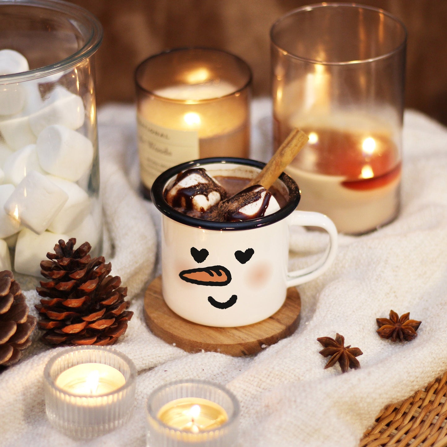 A snowman mug with In Love face rests on a wooden coaster and ivory blanket, surrounded by candle lights, marshmallow, and pine corns. The mug is filled with hot chocolate, marshmallows on top and cinnamon stick.