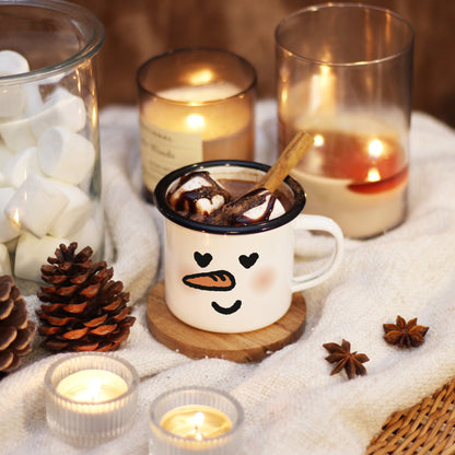 A snowman mug with In Love face rests on a wooden coaster and ivory blanket, surrounded by candle lights, marshmallow, and pine corns. The mug is filled with hot chocolate, marshmallows on top and cinnamon stick.