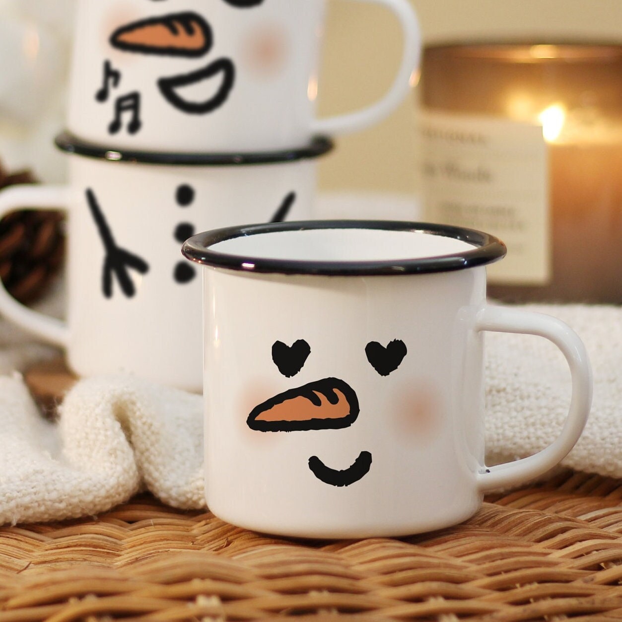 An In Love snowman mug with hand-drawn illustration of heart-shaped eyes, carrot nose, soft blushes, and a smiling mouth, rests on a rattan table with candles on the back.